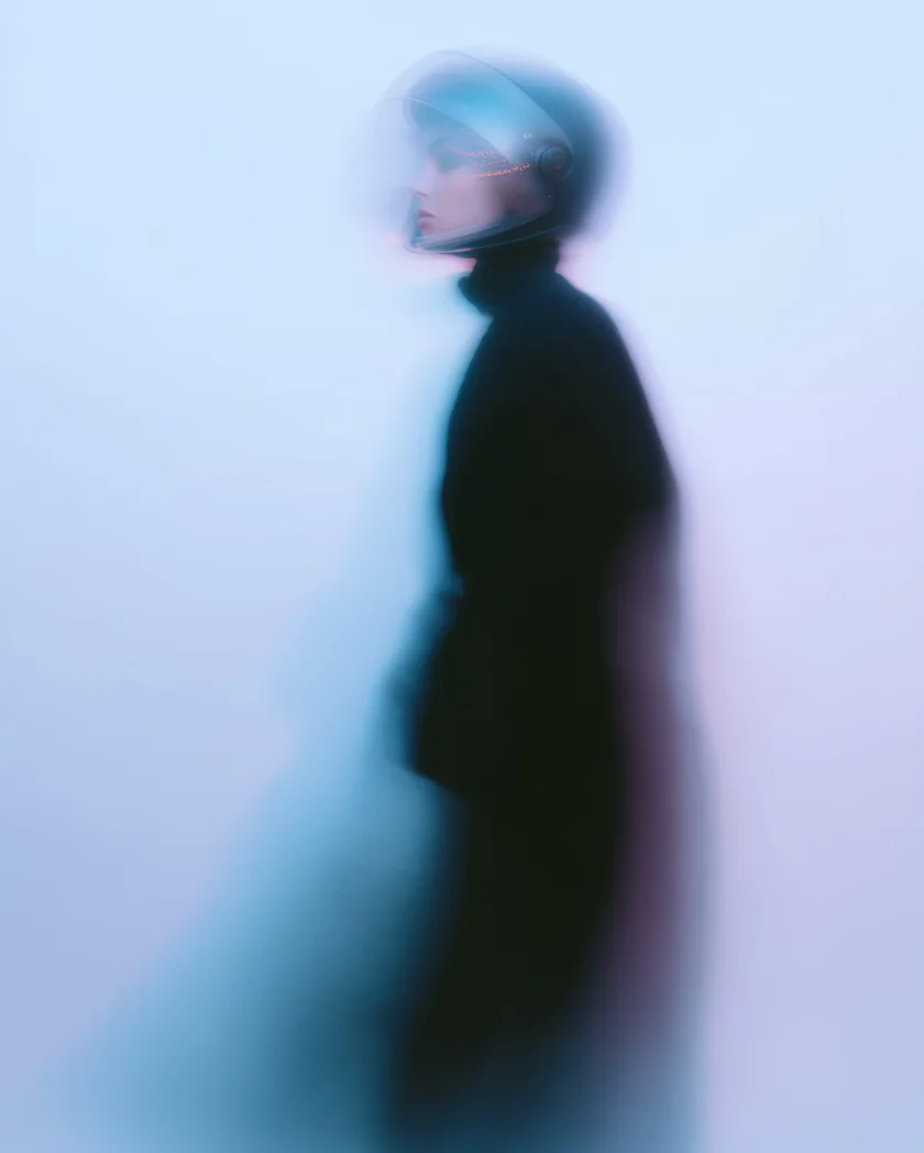 Blurred side profile of a person wearing a futuristic astronaut helmet and dark clothing against a soft blue background.